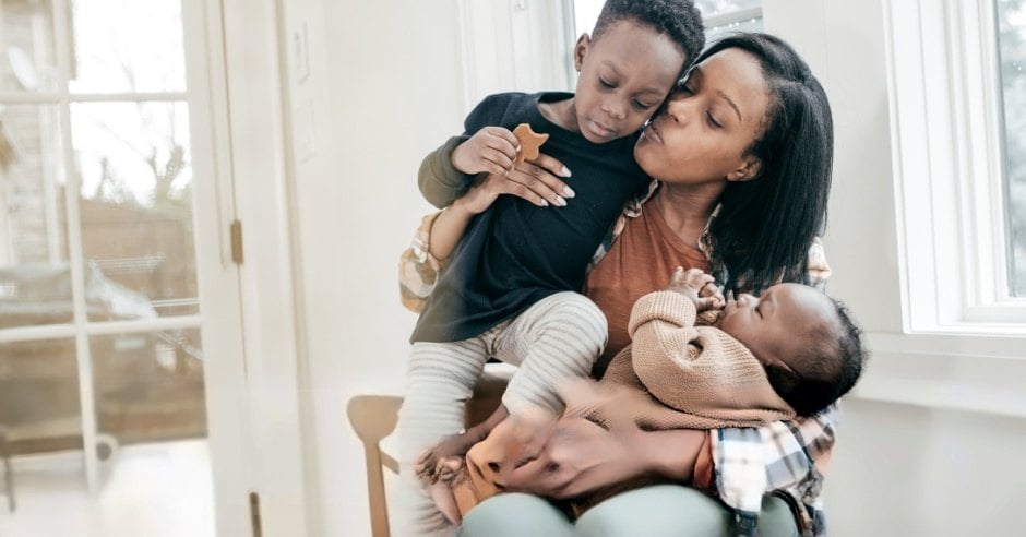 Mother kissing her son while holding her sleeping baby by a bright window